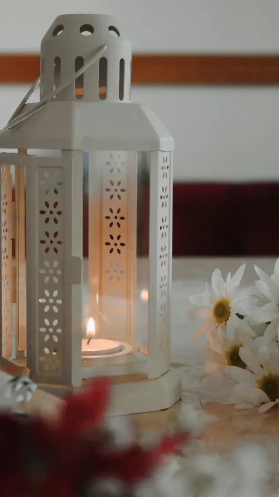 white decorative metal lantern with glowing candle inside beside white daisy flowers on table