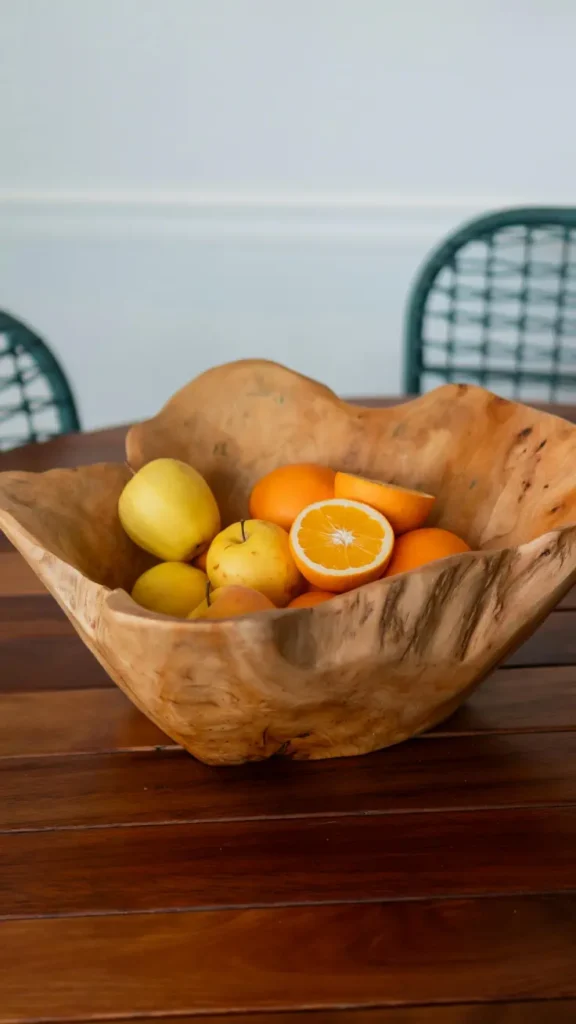 large rustic carved wooden fruit bowl filled with oranges and yellow apples on dark wood dining table with green chairs