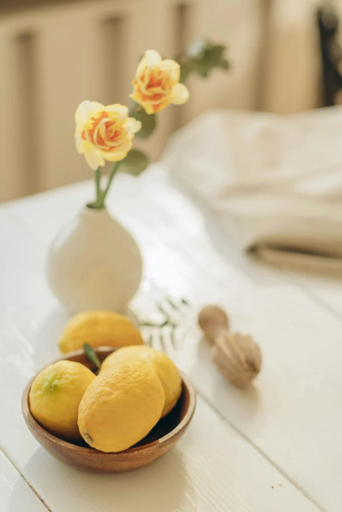 wooden bowl filled with bright yellow lemons on white kitchen table with small white vase and yellow roses in background
