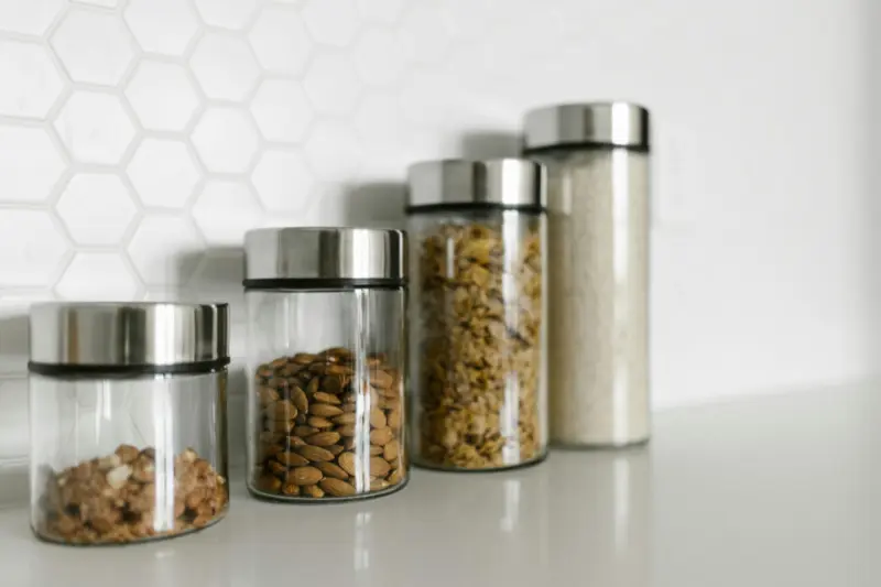 four matching glass jars with silver lids filled with nuts and grains on white kitchen counter for organized storage
