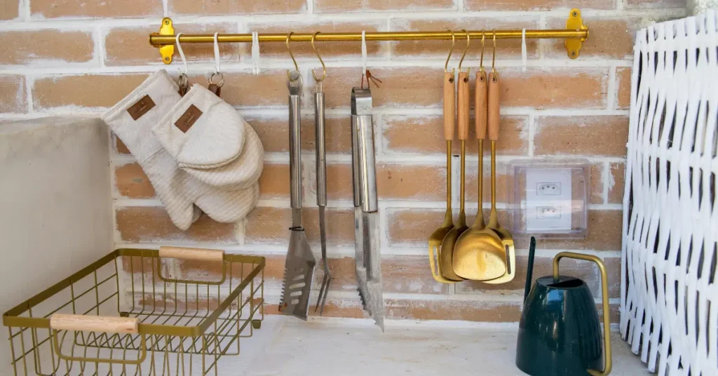 gold and stainless steel grill tools hanging on a brass wall rail with oven mitts beside them against a brick wall in an outdoor kitchen