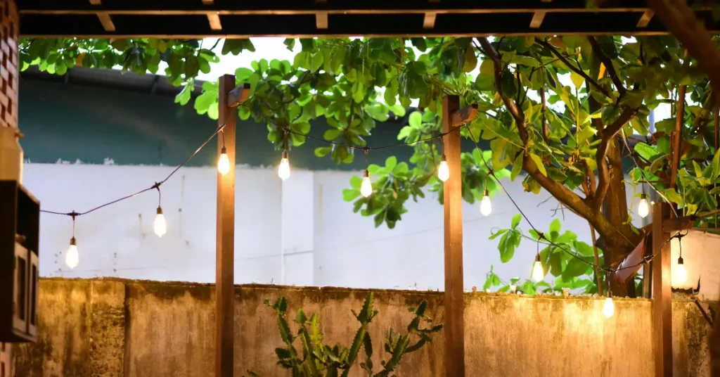 Edison bulb string lights hanging between wooden pergola posts above an outdoor patio area with lush green trees at night