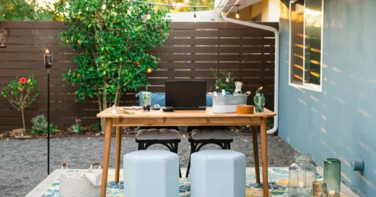 A stylish outdoor dining and workspace setup on a backyard patio, featuring a wooden table, light blue ottomans, string lights, and lush greenery, showcasing stunning outdoor kitchen and backyard decor ideas.