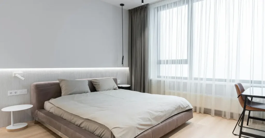 long sheer white curtains hanging from ceiling to floor beside a neatly made bed in a minimal grey bedroom with soft natural light filtering through the fabric