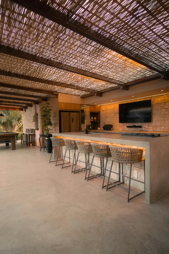 stunning outdoor kitchen and bar area with concrete counter stone wall rattan bar stools and bamboo ceiling all in matching warm neutral earthy tones with amber under counter lighting