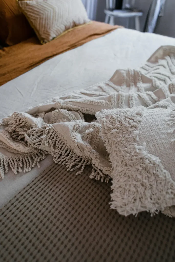 cream and neutral tasselled throw blanket draped casually across a bed with warm rust coloured bedding and chevron pillow visible in the background in a cozy bedroom