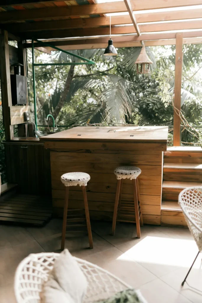 two wooden bar stools with white crochet tops pulled up to a rustic wooden outdoor kitchen island with tropical trees in the background