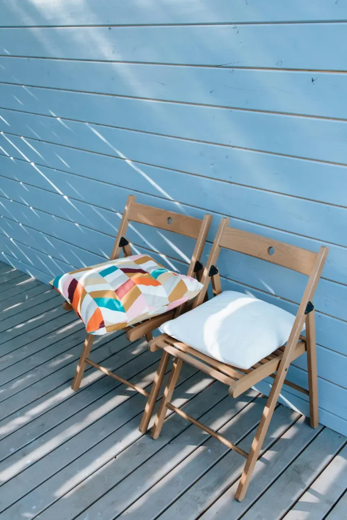two wooden folding chairs on a sunny outdoor deck one with a colorful geometric cushion and one with a white cushion against a light blue painted wall