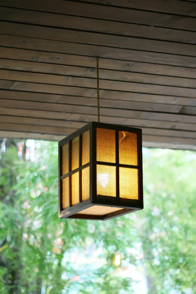 warm glowing square pendant light hanging from a wooden outdoor pergola ceiling with lush green trees in the background