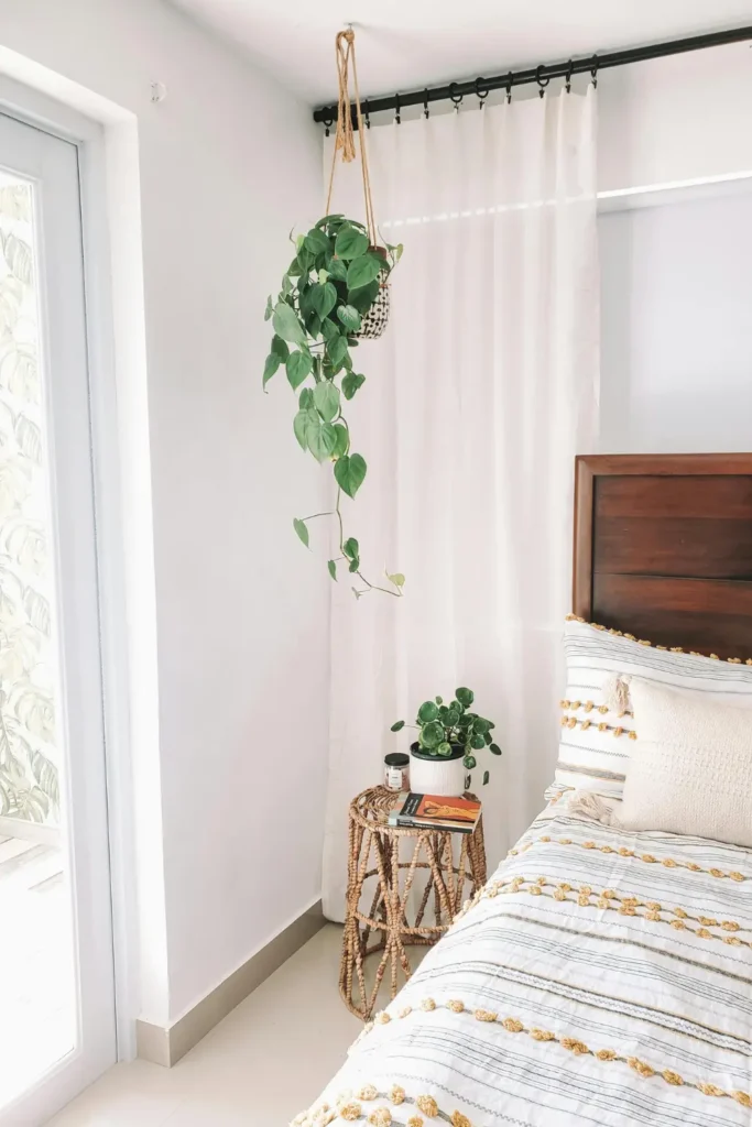 trailing hanging plant in a macrame hanger beside a bed with a small potted plant on a wicker nightstand white curtains and boho striped bedding in a bright bedroom