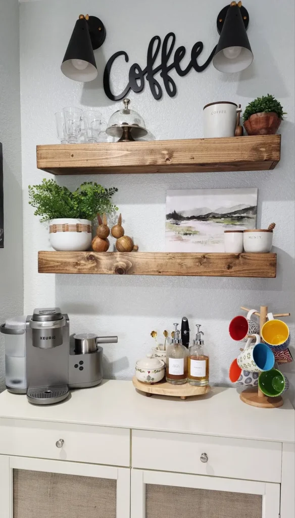 : Two floating wood shelves on a white kitchen wall styled with art prints, books, a plant and mugs hanging on a rail below beside a coffee machine
