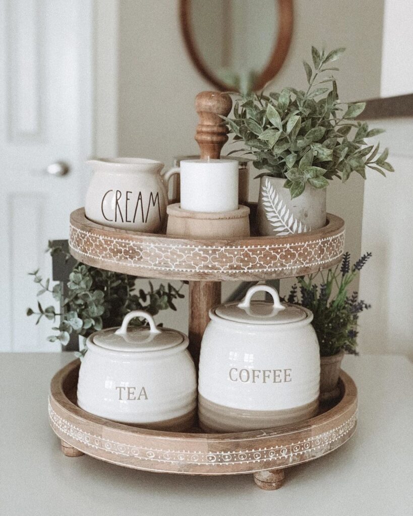  Cream ceramic labeled tea and coffee canisters on a two-tier carved wooden tray stand with small green plants and a cream jug on a kitchen counter