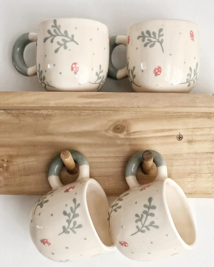 Two matching hand-painted ceramic mugs with mushroom and botanical pattern sitting on a wooden shelf with two more hanging from hooks below