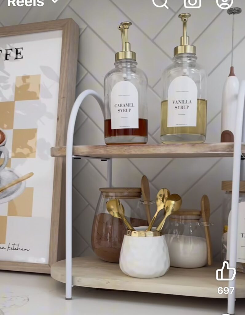 Glass syrup dispensers labeled caramel and vanilla with gold pumps on a small tiered wooden rack beside gold spoons and glass canisters on a kitchen counter