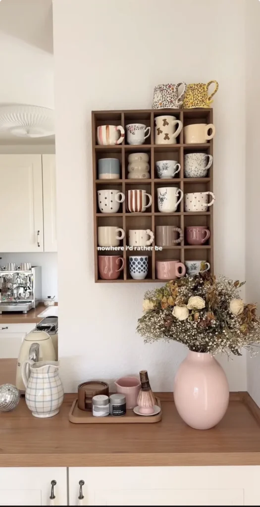 Wall mounted wooden mug display shelf with individual compartments holding a collection of mismatched ceramic mugs in pink, cream, and patterned styles above a styled kitchen counter with a wooden tray and pink vase
