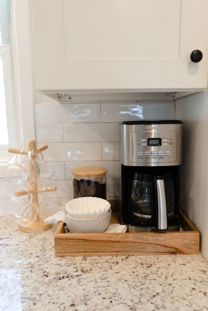 Kitchen coffee station with a flat wooden tray holding a stainless steel coffee maker and glass coffee canister beside a wooden mug tree on a granite counter