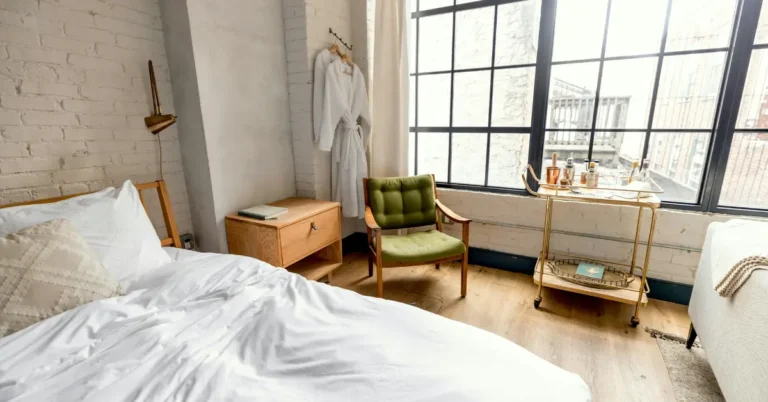 Sunlit loft studio apartment featuring a white unmade bed, green mid-century armchair, and a gold bar cart by large industrial grid windows.