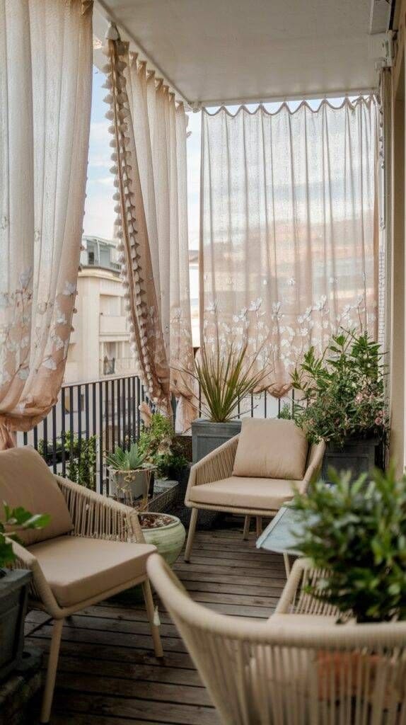 Small apartment balcony with sheer pink floral curtains hanging from the ceiling, rattan chairs, green plants and a wooden deck floor overlooking a city view.