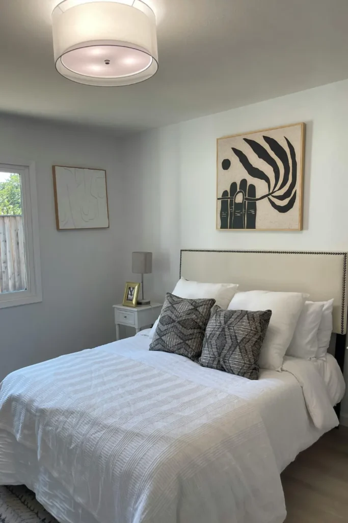 bold black and cream abstract art print in a wooden frame hanging above a neatly made bed with white bedding and dark patterned pillows in a minimal grey bedroom