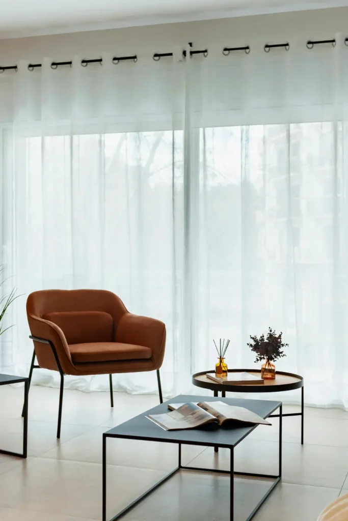 A bright minimal apartment living room with white sheer curtains covering a large window, a rust colored armchair and black coffee tables in soft natural light.