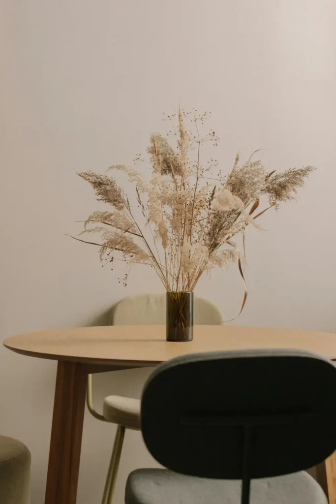 A round wooden dining table with a dark glass vase holding a large bunch of dried pampas grass and wildflowers, soft neutral toned apartment background with green chairs.