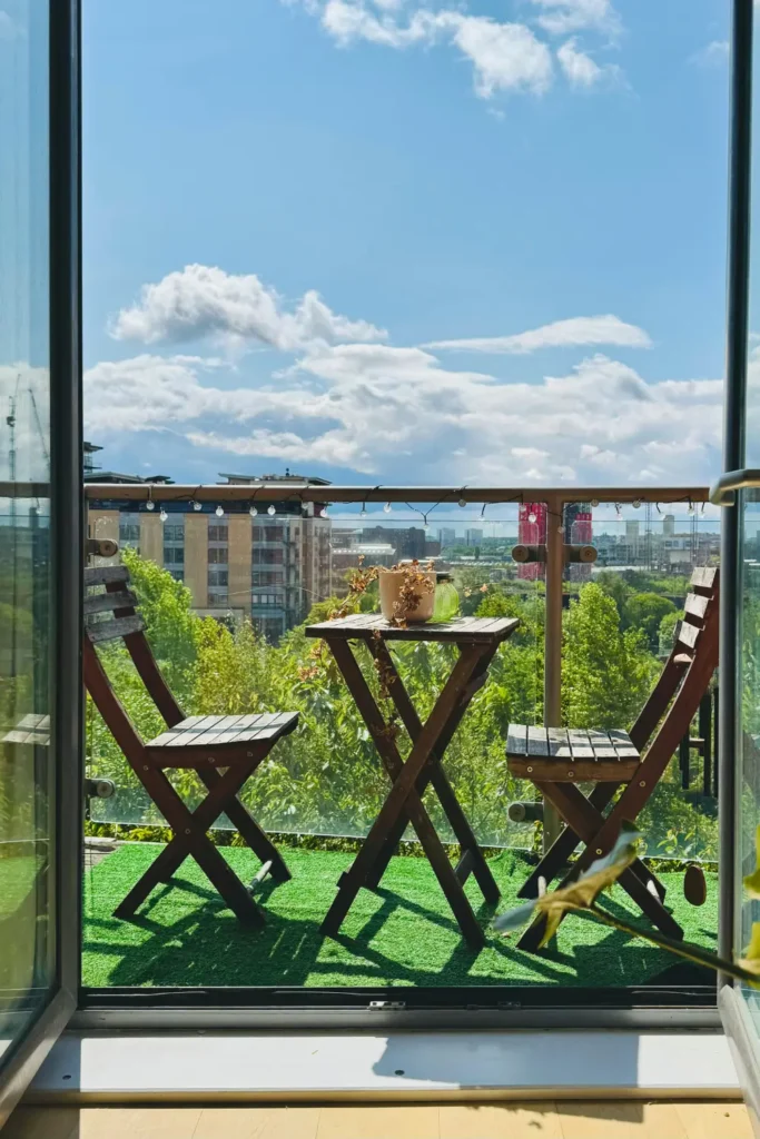 Small apartment balcony with wooden folding chairs and a bistro table facing a green city view through a glass railing on a sunny day.