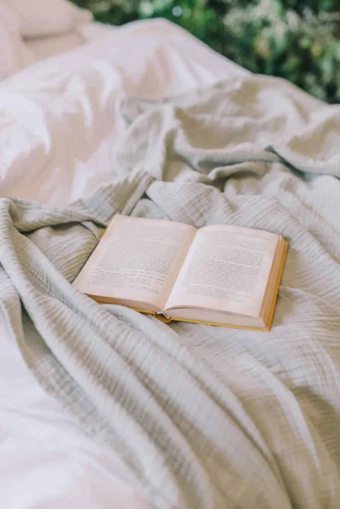 A close up of soft light grey cotton bedding with a yellow book open on top, natural light and greenery visible in the background.