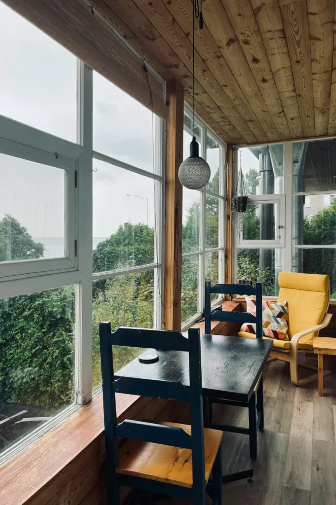 Enclosed apartment balcony with a wooden ceiling, black table and chairs, a yellow armchair and floor to ceiling windows overlooking green trees.