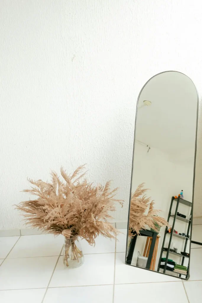 tall arched full length mirror with a thin dark frame leaning against a white wall beside a large dried pampas grass arrangement on a white tiled floor