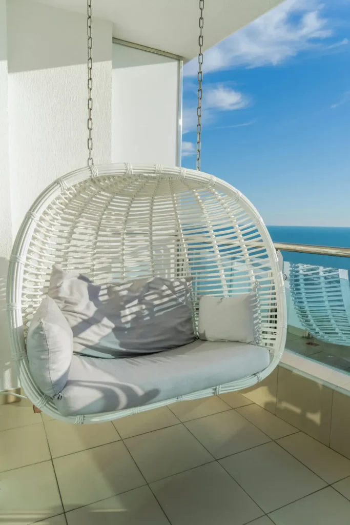 White rattan hanging egg chair with grey cushions on a bright apartment balcony overlooking the ocean and blue sky.
