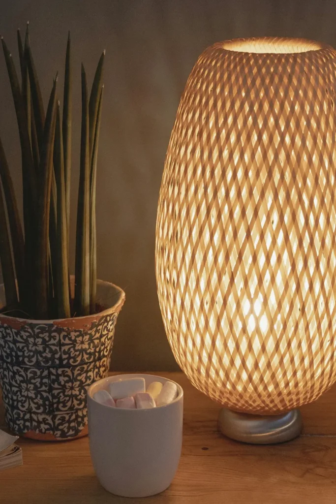 A glowing woven rattan table lamp on a wooden nightstand next to a small potted snake plant and a white ceramic cup in a dim apartment bedroom.