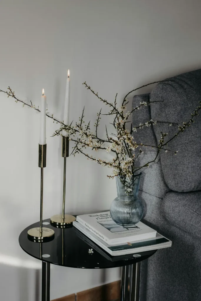 A black round side table next to a grey sofa styled with gold candlesticks, white taper candles, a blue glass vase with flowering branches and stacked books.