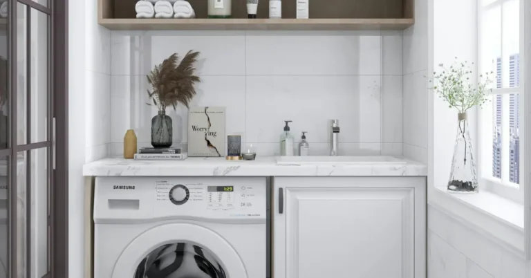Small apartment laundry room with a marble countertop above a Samsung washing machine, white cabinets, open wood shelving with folded towels and products, and a styled vase with dried pampas grass.
