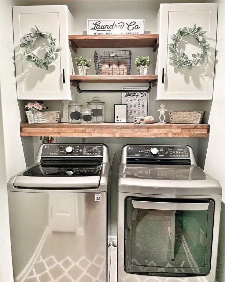 Small laundry room with rustic wood open shelving holding labeled glass canisters, woven baskets and greenery above a side by side washer and dryer with white cabinets.