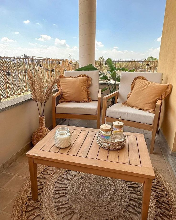 Small apartment balcony with a wooden table styled with a candle and a small tray, two wooden chairs with orange cushions, a jute rug and a bamboo privacy screen in the background.