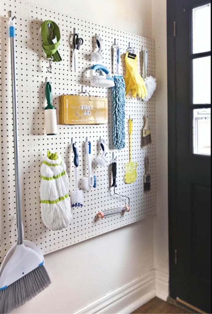 White pegboard organizing cleaning tools including a broom, scrub brushes, rubber gloves, and a first aid box on a laundry room wall