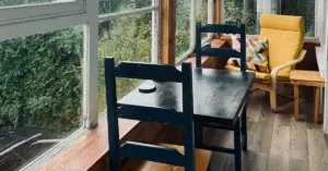 Enclosed apartment balcony with a black wooden table and chairs, a yellow armchair and floor to ceiling windows overlooking lush green trees.