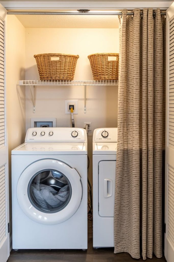 Small closet laundry room with a striped tension-rod curtain pulled open beside a washer and dryer, wicker baskets on a wire shelf above