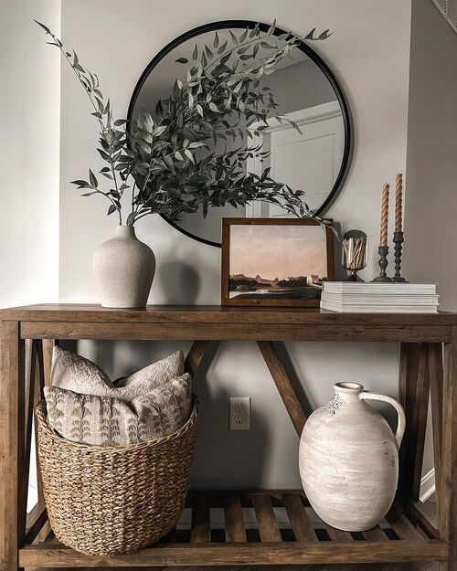 Small apartment entryway with a narrow wood console table, round black mirror, white ceramic vase with greenery, and woven basket underneath