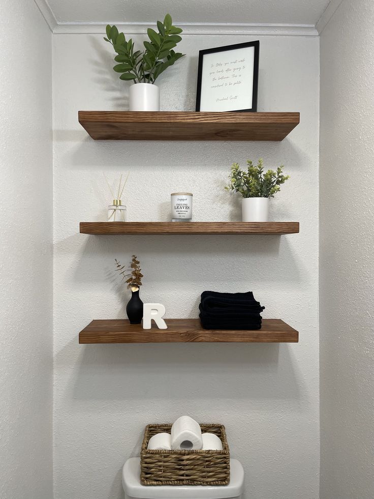 Small apartment bathroom with three stacked wood floating shelves above the toilet styled with a plant, framed quote, candle, diffuser, folded black towels and a small vase, with a woven basket of toilet paper on the tank