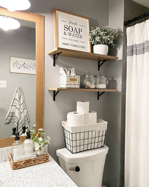 Wood floating shelves above toilet with glass apothecary jars holding cotton balls and Q-tips, framed soap and water print and wire basket with toilet paper rolls on toilet tank