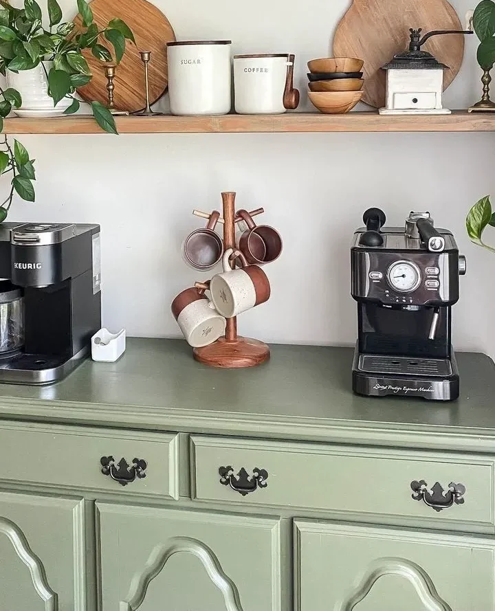 A copper countertop mug tree holding white mugs standing between a Keurig and espresso machine on a green dresser used as a small apartment coffee corner