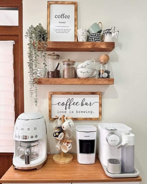 A white SMEG coffee machine setup with a coffee bar love is brewing sign mounted above floating wooden shelves holding mugs and canisters in a small kitchen