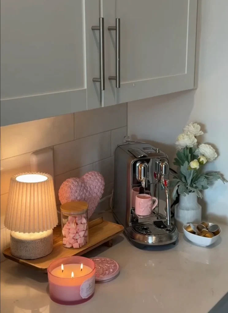 A cozy small apartment coffee corner with a small glowing pleated lamp next to a silver Nespresso machine and a lit candle and fresh white flowers on the counter