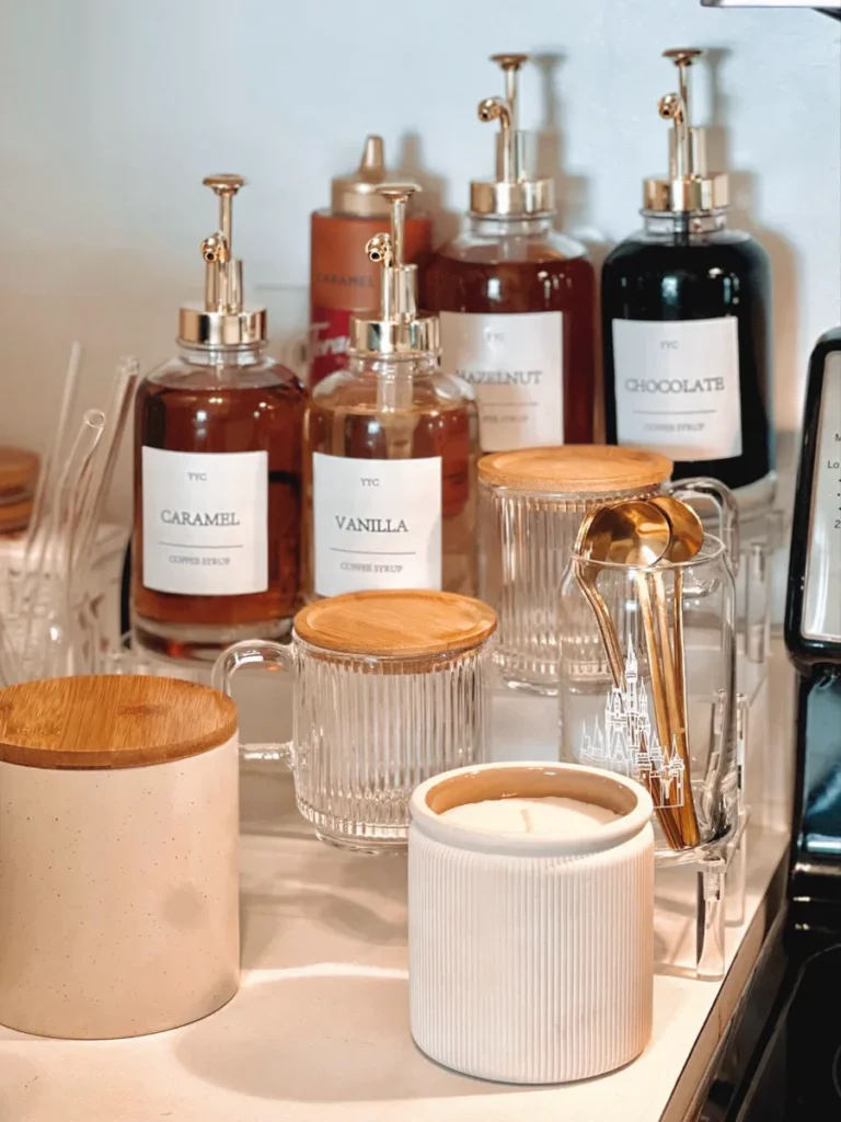 Labeled coffee syrup bottles in caramel vanilla hazelnut and chocolate lined up on a coffee corner counter with a white ribbed ceramic candle and glass jars with bamboo lids in front