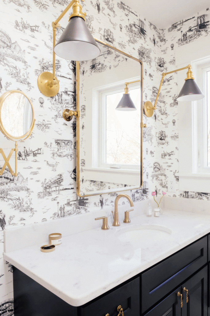 Small apartment bathroom with two brass and black wall sconces flanking a large rectangular brass mirror over a white marble vanity with black and white toile wallpaper