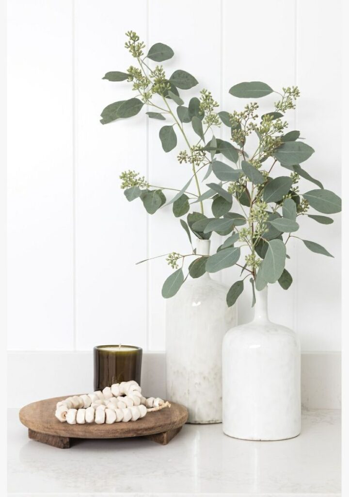 Small apartment bathroom counter styled with a round rustic wooden riser, green candle, white beaded garland, and a white ceramic vase with eucalyptus stems against a white paneled wall