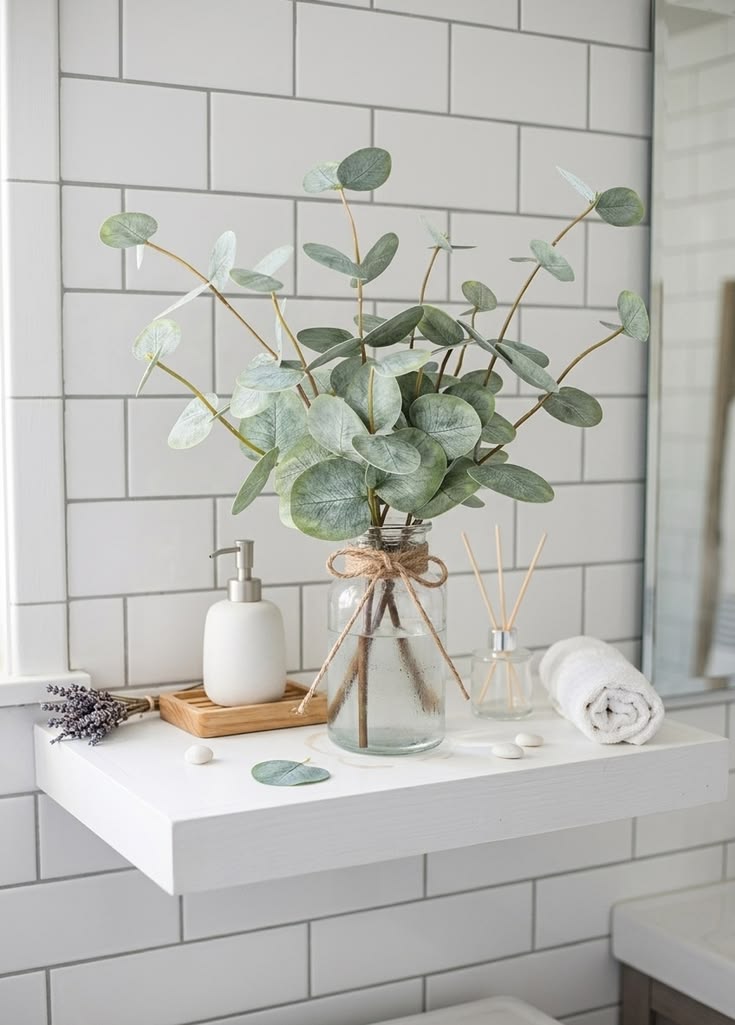 Small apartment bathroom counter with a glass jar of faux eucalyptus stems tied with twine, white soap dispenser, reed diffuser, and rolled white towel against subway tile