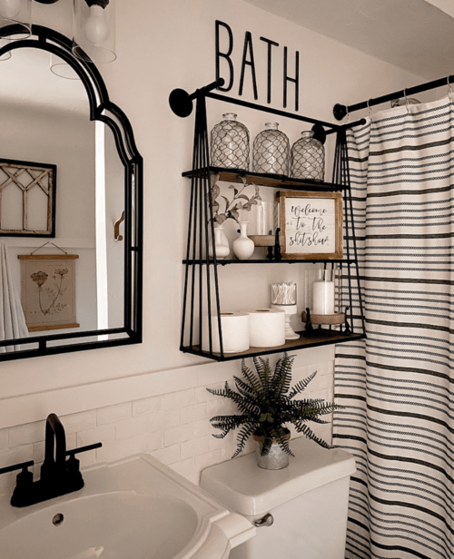 Small apartment bathroom with black two-tier metal floating shelves above toilet styled with glass jars, white vases and a small fern plant, black arch mirror on the wall, black and white striped shower curtain and a BATH sign above the shelves