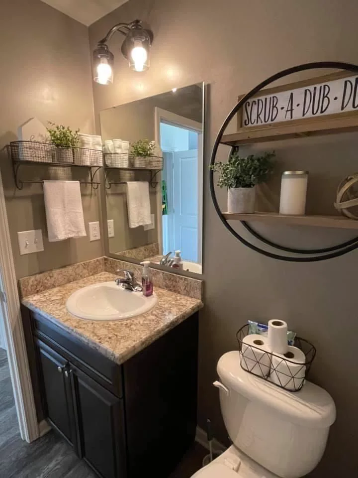 Dark metal circular shelf and rectangular mirror on bathroom wall above toilet area with wire basket holding toilet paper on toilet tank